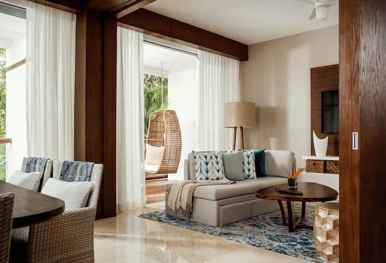 A one-bedroom deluxe room at the Vidanta Riviera Maya resort, featuring a living area with a sofa bed, a small dining table, and large sliding glass doors that open to a balcony with a hanging wicker chair.