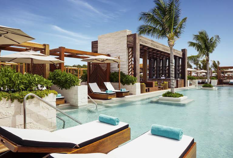 A large swimming pool at the Beach Club at Vidanta Riviera Maya, surrounded by palm trees, sun loungers, and umbrellas.