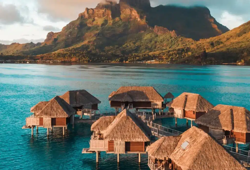 Overwater bungalows with thatched roofs at Four Seasons Resort Bora Bora, with a backdrop of a lush mountain and a clear blue lagoon.