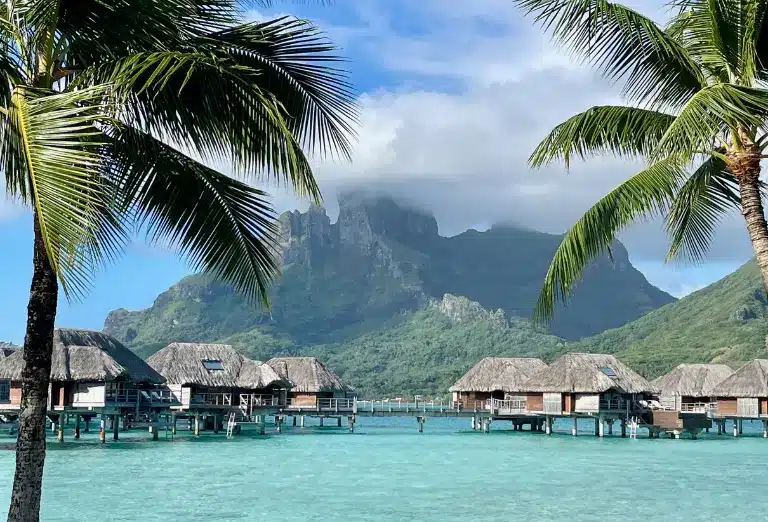 Scenic view of the overwater bungalows at Four Seasons Resort Bora Bora with Mount Otemanu shrouded in clouds, framed by tropical palm trees, capturing the essence of a serene island retreat.