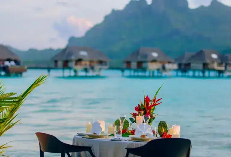 Intimate dinner table set for two with candles and a floral arrangement at the Four Seasons Bora Bora, with overwater villas and mountain scenery in the background.