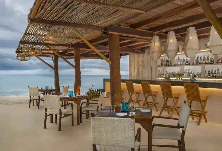Elegant beachfront dining at the Coral Bar in Viceroy Riviera Maya, with an open-air design, woven light fixtures, and a tranquil view of the ocean, providing an ideal spot for a refreshing cocktail or a gourmet meal.
