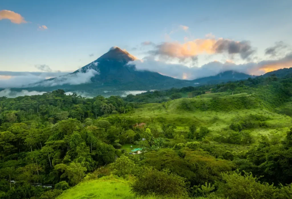 Arenal Volcano in Costa Rica is surrounded by dense green rainforest under a blue sky with clouds encircling the volcano.