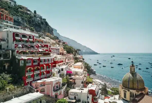Le Sirenuse resort in Positano, Italy, overlooking the Amalfi Coast with colorful hillside buildings, church dome, and boats on the turquoise sea.