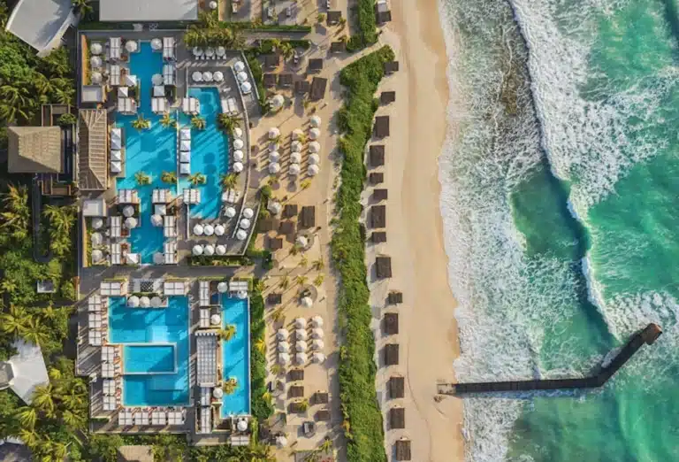 An aerial view of the Vidanta Riviera Maya resort, showing a complex of turquoise swimming pools, a sandy beach with rows of cabanas and umbrellas, and the turquoise waters of the Caribbean Sea with a long pier extending into the water.