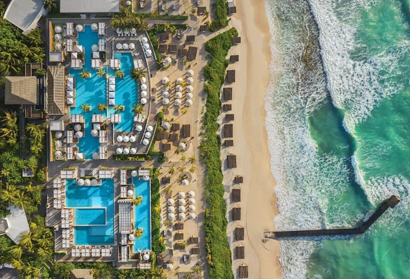An aerial view of the Vidanta Riviera Maya resort, showing a complex of turquoise swimming pools, a sandy beach with rows of cabanas and umbrellas, and the turquoise waters of the Caribbean Sea with a long pier extending into the water.