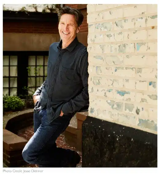 A portrait of actor and travel writer Andrew McCarthy, smiling and leaning against a white brick wall in a garden setting.