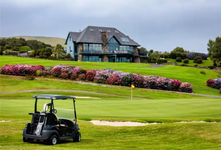 A black golf cart parked on a lush, rolling green course in Ireland near Sheen Falls Lodge, with a modern stone clubhouse and vibrant pink hydrangeas in the background.