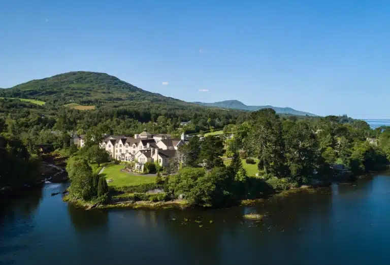 An aerial view of the luxurious Sheen Falls Lodge in Ireland, a sprawling yellow country house hotel nestled between lush green forests and the calm waters of Kenmare Bay, as described by Andrew McCarthy.