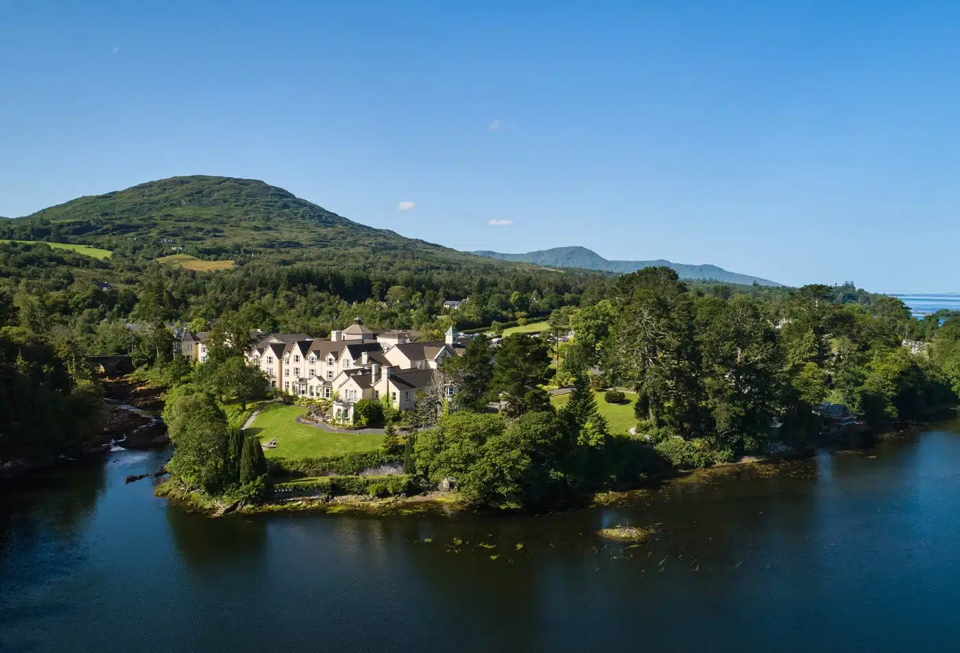 An aerial view of the luxurious Sheen Falls Lodge in Ireland, a sprawling yellow country house hotel nestled between lush green forests and the calm waters of Kenmare Bay, as described by Andrew McCarthy.