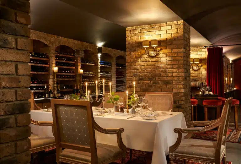 An atmospheric candlelit dinner setting within the rustic brick-vaulted wine cellar at Sheen Falls Lodge, featuring a white linen table surrounded by thousands of vintage bottles.