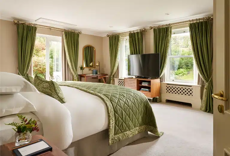 A bright and spacious luxury bedroom at Sheen Falls Lodge, featuring green botanical accents, a plush king-sized bed, and large windows overlooking the lush Irish gardens of the Kenmare Bay area.